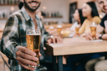 selective focus of man holding glass of light beer while sitting in pub with friends