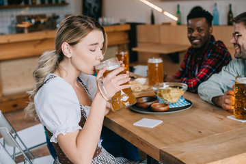 selective focus of young woman in traditional german costume drinking beer near multicultural friends