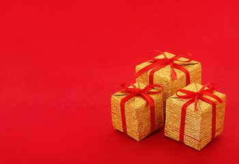 Stack of small golden gift box with red ribbon and bow on red background, with empty place for text on the left. Close up.