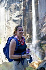 Young woman stands in-front of a waterfall in Yosemite National Park, with her water bottle for hydration. 