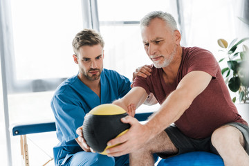 Obraz premium doctor sitting near mature patient exercising on fitness ball