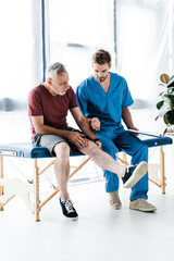 Fototapeta na wymiar bearded mature patient touching leg near doctor holding clipboard in clinic