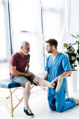 handsome doctor touching leg and looking at bearded mature patient