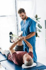 handsome doctor holding leg of mature patient exercising with elastics