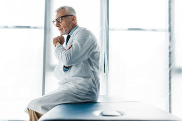 Fototapeta premium thoughtful doctor in glasses and white coat sitting on massage table and touching face