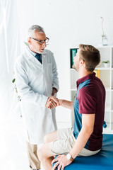 happy doctor in glasses shaking hands with young man in clinic