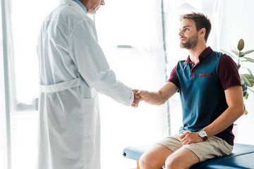 Obraz premium cropped view of doctor in white coat shaking hands with handsome man