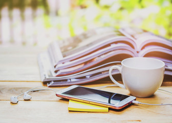 close up of smart phone and yellow note pad  over blurred stack of magazine on wooden table against sun light, nature background