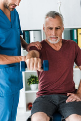 Obraz premium cropped view of bearded doctor standing near middle aged man exercising with dumbbell