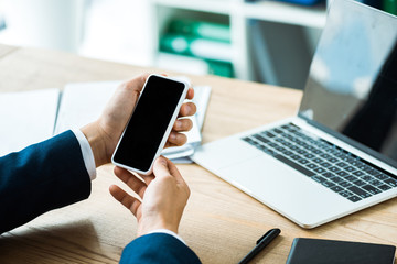 cropped view of businessman holding smartphone with blank screen near laptop on table