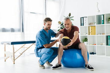 mature patient exercising on fitness ball near doctor