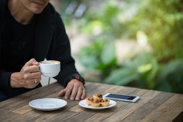 Man holding a cup of coffee with pastry and smart phone on wooden table outdoors.