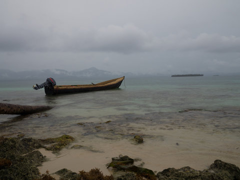 Small boat anchored next to a palm log on a rainy day, mountain range on the background