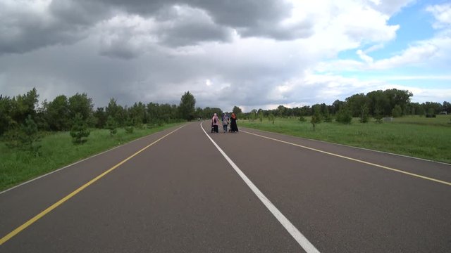 Women in hijabs with prams. Asphalt walkway in the park. View from the back. Road marking lines. Green trees. Rain clouds in the sky.