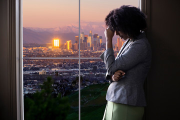 Black female businesswoman looking worried or tired by an office window with a view of downtown Los...