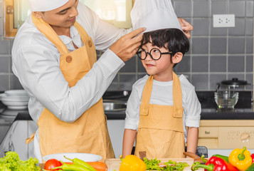 Father Wear chef hat for son boy holdind wooden spoon cooking salad food with vegetable holding tomatoes and carrots, bell peppers on plate for happy family cook food enjoyment lifestyle kitchen