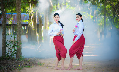 Two beautiful asian women in white shirt and red loincloth holding pink lotus flowers. Two young asian women with sarong cloth decorated with silver belt according to Thai culture, Thailand. - obrazy, fototapety, plakaty