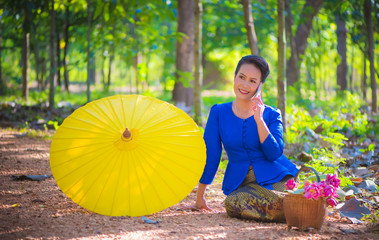 An Asian woman in blue shirt is calling her mobile phone with Yellow umbrella,  A woman who is happy smile talking on the smartphone with pink lotus flowers in bamboo basket and forest background.