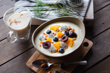 Yogurt with cherries, blueberries, pineapples, pitayas, nuts and almond milk on a brown table.