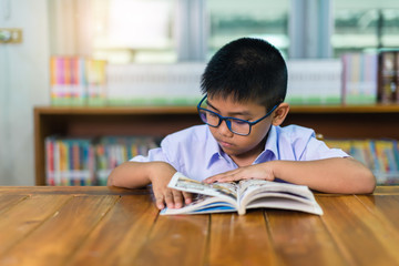 A cute Asian elementary school boy wearing blue glasses in a white school uniform is sitting, enjoying , reading comics in the library.