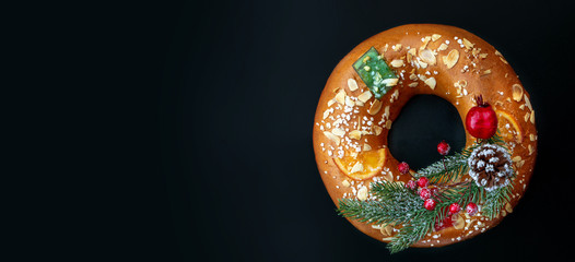 Christmas round fruit cake decorated with fir tree branch, glazed fruits and nuts over  black background. Roscon de reyes. King cake Top view. Copy space