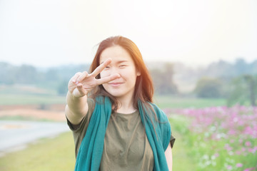 Portrait of young asian woman looking at camera with smile and showing peace sign with fingers with cosmos field in background.