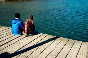 Young couples in winter clothes sitting on wood bridge, relax and enjoy remarkable scenery turquoise Wakatipu Lake, best popular location for tourists and photographers in Glenorchy, New Zealand.