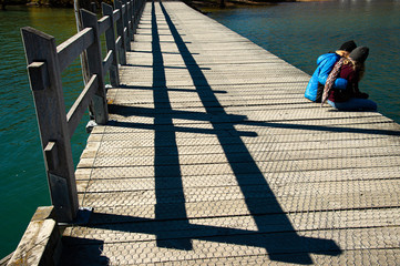 Young couples traveler sitting on wood bridge, relax and enjoy remarkable scenery turquoise Wakatipu Lake, best popular location for tourists and photographers in Glenorchy, South New Zealand.