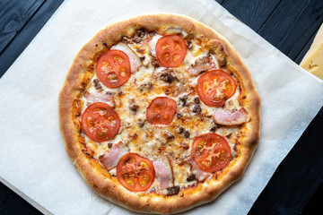 Top view tasty Pizza on craft paper on a black background. Pizza with ham, tomatoes and mushrooms. Italian Cuisine. Food photo background