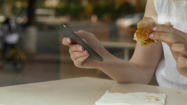 Woman browse pages using smartphone, sit at cheap fast food restaurant, slow motion shot through window glass. She scroll news feed or images and hold bitten piece of burger in other hand