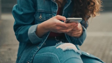 Close up of the smartphone being in hands of the Caucasian young girl who typing and texting a message while chatting in social media and sitting outdoors.