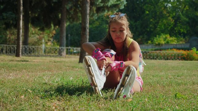 Young schoolgirl sitting on the grass in a city park putting on her rollerblades - closing up the clasp. 4k video