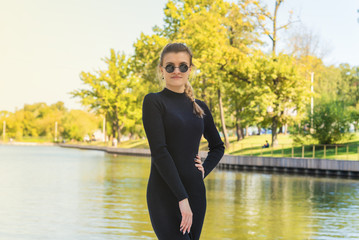 Young attractive slim woman in a tight-fitting black dress and sunglasses. Autumn park with a pond in the background. Beautiful fashionable woman on a park background.