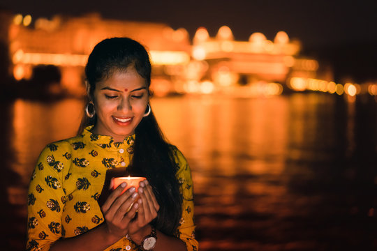 Young beautiful Indian woman holding a lit candle for Indian Festival Diwali at Ambrai Ghat in front of Picola Lake and City Palace, Udaipur, Rajasthan, India. 