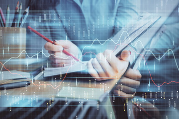 A woman hands writing information about stock market in notepad. Forex chart holograms in front. Concept of research. Multi exposure