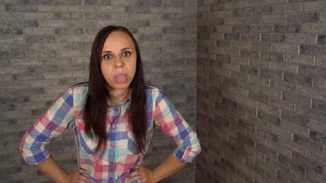 A beautiful young woman in a plaid shirt shows with insult her tongue looking at camera on a brick background.