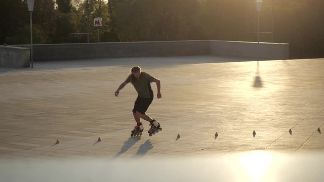 Young long-haired man roller skater is dancing between cones in the evening in a city park at sunset. Freestyle slalom Roller skating between cones.