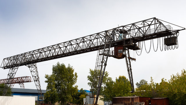large metal gantry crane at a construction site, in the background industrial warehouses for storing goods. Type of bearing metal structures of gantry crane against the blue sky