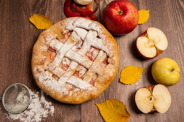 Home cooking. Delicious Apple pie, powdered sugar, red apples and yellow autumn leaves on wooden background.