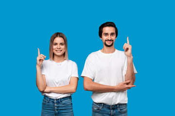 Happy couple in white t-shirts and jeans pointing up with fingers standing isolated over blue background.
