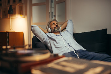 mid aged man listening music with headphones on phonograph, relaxed in sofa at his home - obrazy, fototapety, plakaty