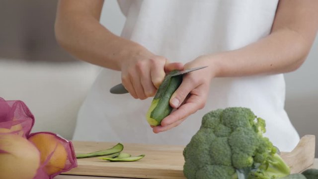 Close-up of hands of young slim woman peeling cucumber with the sharp knife at the table in the kitchen. Concept of healthy food. Fruit and vegetables lying in the foreground