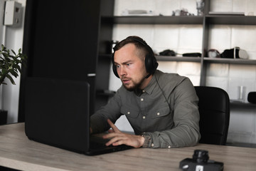 Confused or angry businessman or student in a shirt sitting against monitor of computer in headphones and speaking with somebody via internet. Working on a pc at a table in the office.