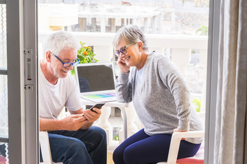 couple of seniors smiling and looking at the phone with coffe and laptop in the terrace - outoor...
