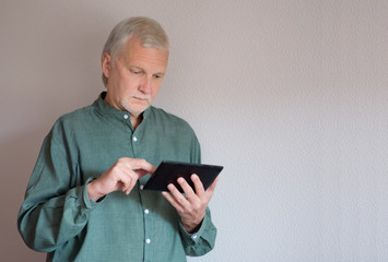 Elderly man dressed in a green shirt stands and holds a tablet using his fingers to browse the Internet.