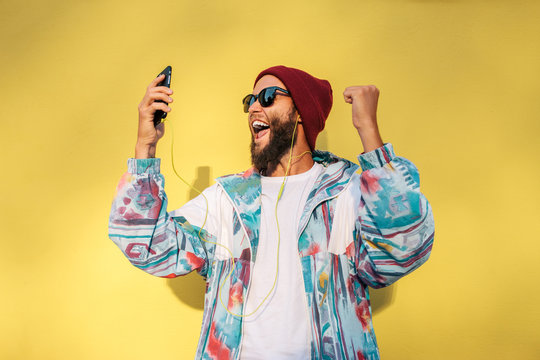 Stylish young hipster man with beard in red hat and a retro jacket of 90s on yellow background dancing and singing while he listens to the music in his smartphone. Relax. love live
