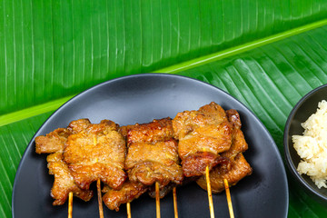 Grilled pork with sticky rice or Khao Niew Moo Yang on black ceramic plate, local Thai food on fresh banana green leaf.