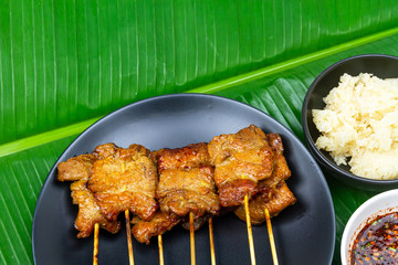 Grilled pork with sticky rice or Khao Niew Moo Yang on black ceramic plate and spicy sauce , local Thai food on fresh banana green leaf.