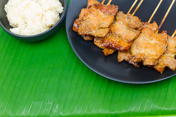 Grilled pork with sticky rice or Khao Niew Moo Yang on black ceramic plate, local Thai food on fresh banana green leaf.