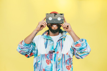 Stylish young hipster man with beard in red hat, sunglasses and a retro jacket of 90s with VHS cassette on yellow background.